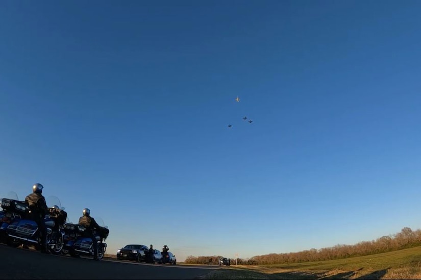 Motorcyclists on a road watch as a plane moves ahead of three others on a cloudless day.