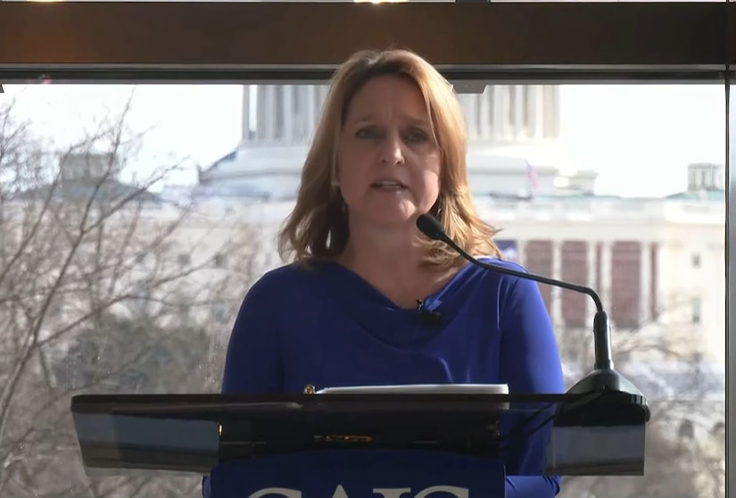 A woman is standing indoors at a podium delivering remarks into a microphone. The U.S. Capitol building is outside in the background behind her.