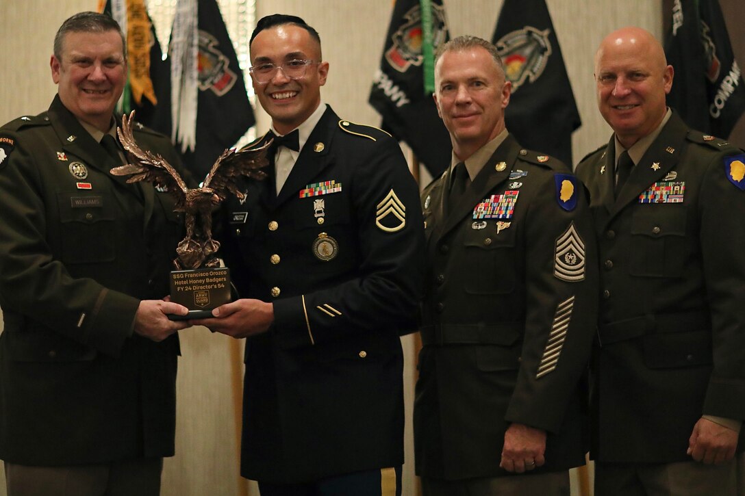 Four soldiers smile in front of standing flags while two hold a trophy.