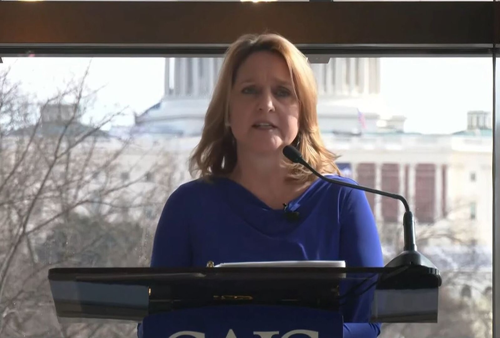 A woman is standing indoors at a podium delivering remarks into a microphone. The U.S. Capitol building is outside in the background behind her.