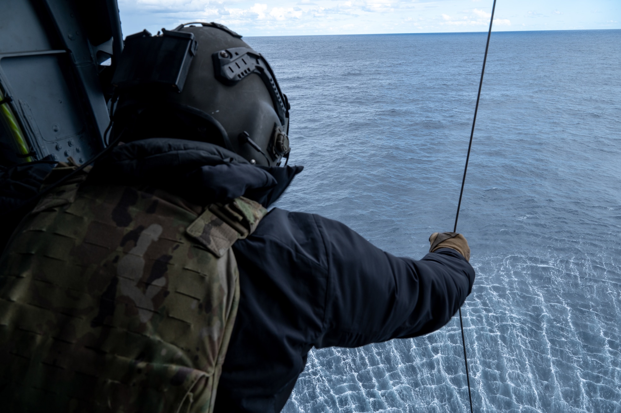 An Airman lowers a hoist form a helicopter over an ocean.