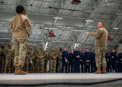 Photo of a U.S. Air Force Airman in the foreground briefing hundreds of service members in the background inside a hangar.