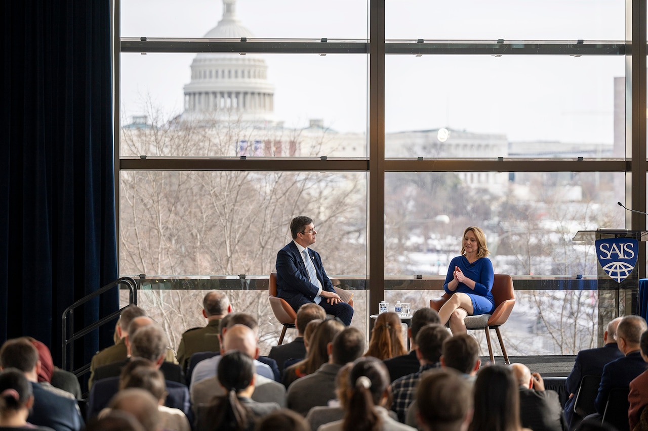 Two people chat on a stage in front of an audience while seated in front of glass windows overlooking the U.S. Capital building.