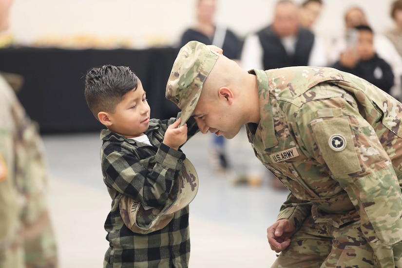 A child adjusts a soldier’s hat as others watch in the blurred background.