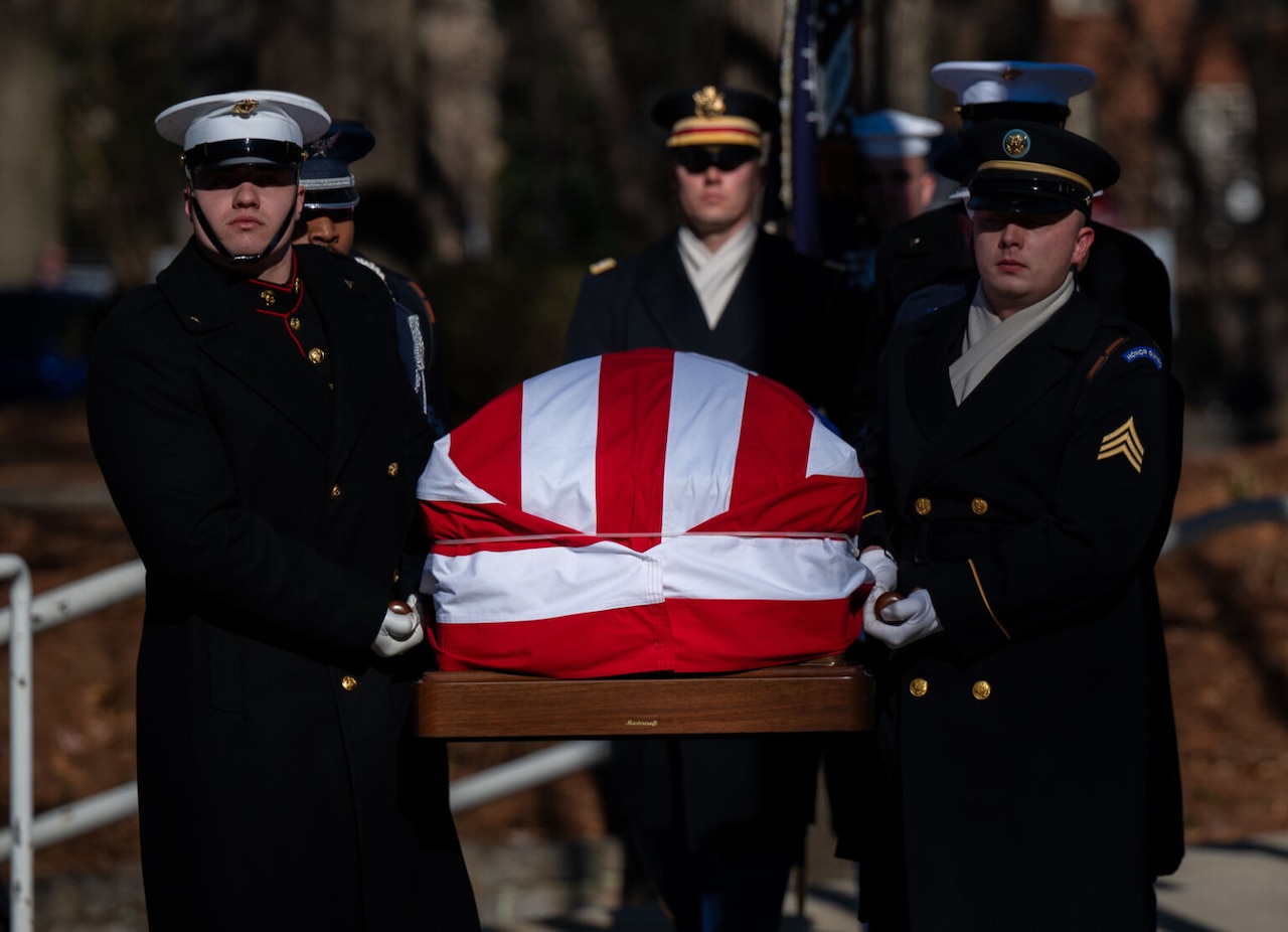 Service members in dress uniforms carry a flag-draped casket during a funeral.