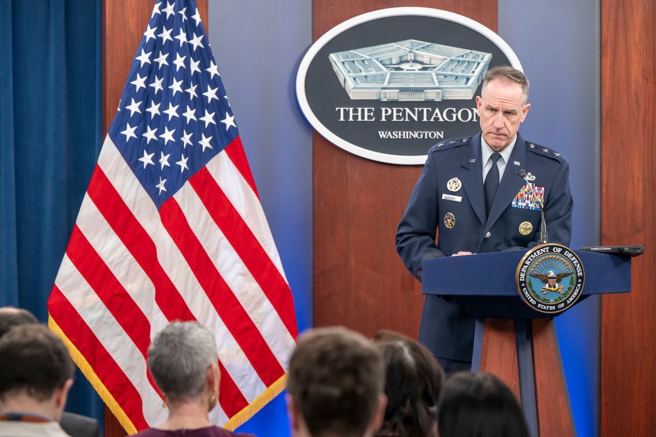 A man in a dress military uniform stands at a podium addressing audience members with their backs to the camera. Behind the man is a wall-mounted sign displaying a five-sided building that reads “The Pentagon.” A U.S. flag is to the man’s right.
