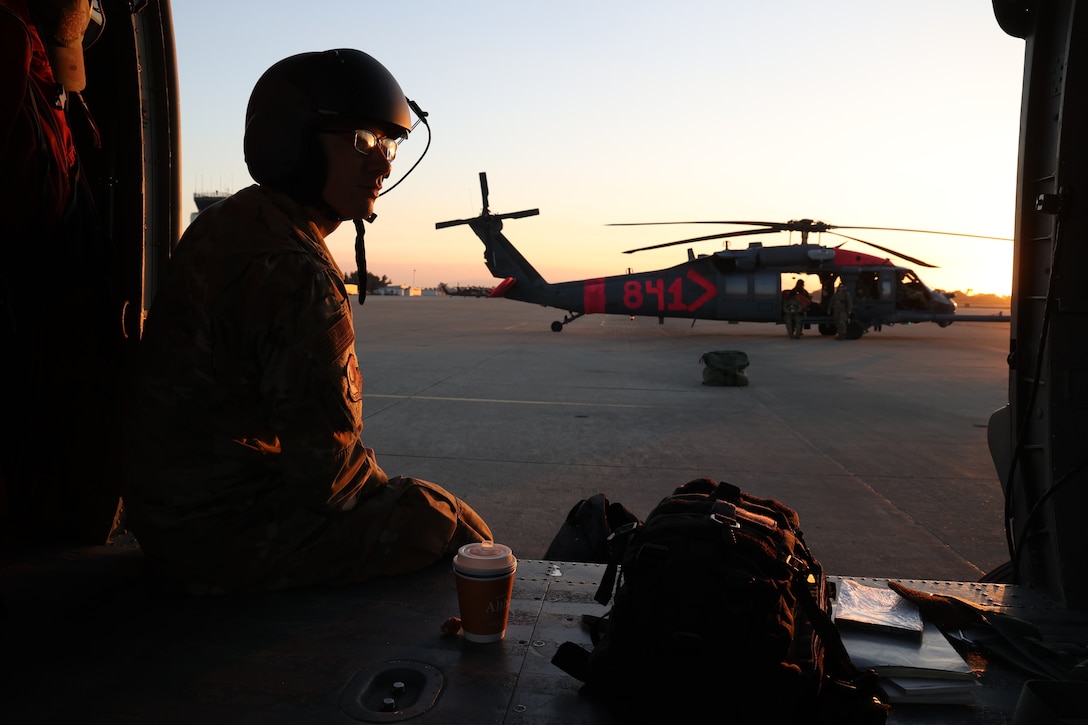 A silhouetted man in a helmet is sitting inside a helicopter with his legs hanging out of the side. Another military helicopter is in the background.