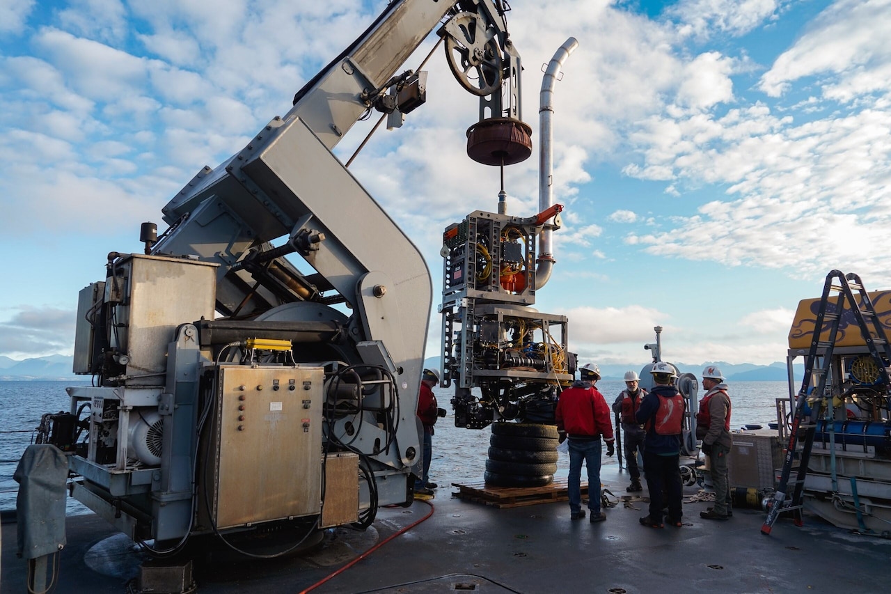 A large piece of machinery dangles from a crane on a ship as five men stand nearby.