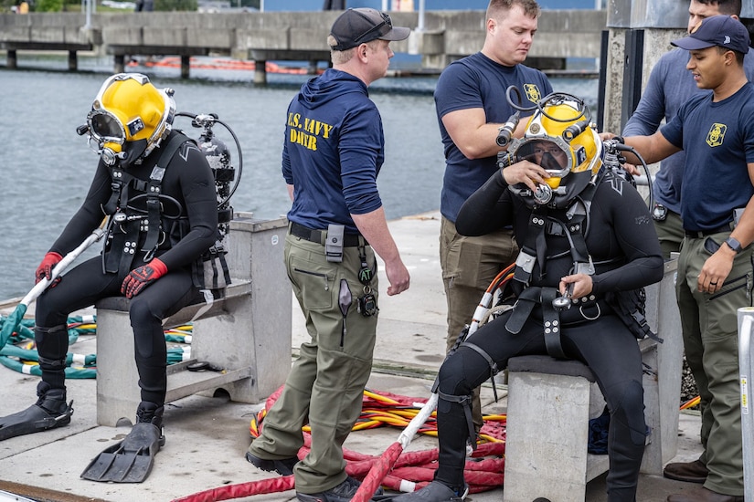 Four men work on equipment attached to a diving helmet on a man dressed in a wetsuit and diving gear. Another diver sits nearby.