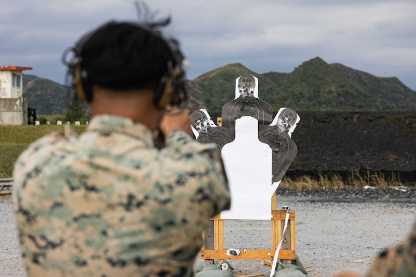 A Marine aims and fires a pistol at a target while wearing ear protection outdoors during a cloudy day.