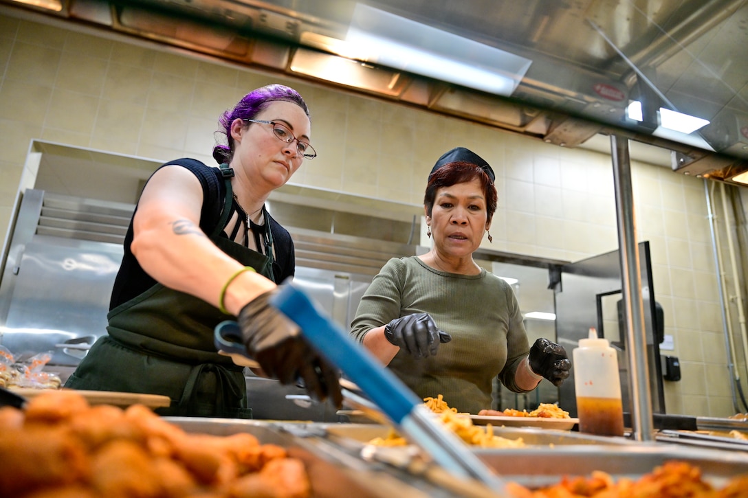 Two women stand behind a food service counter while one scoops food.
