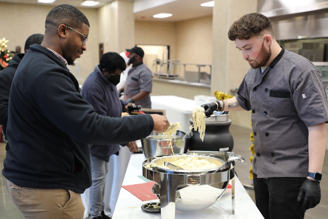 A man in a grey food service uniform serves fettucine noodles to a customer.