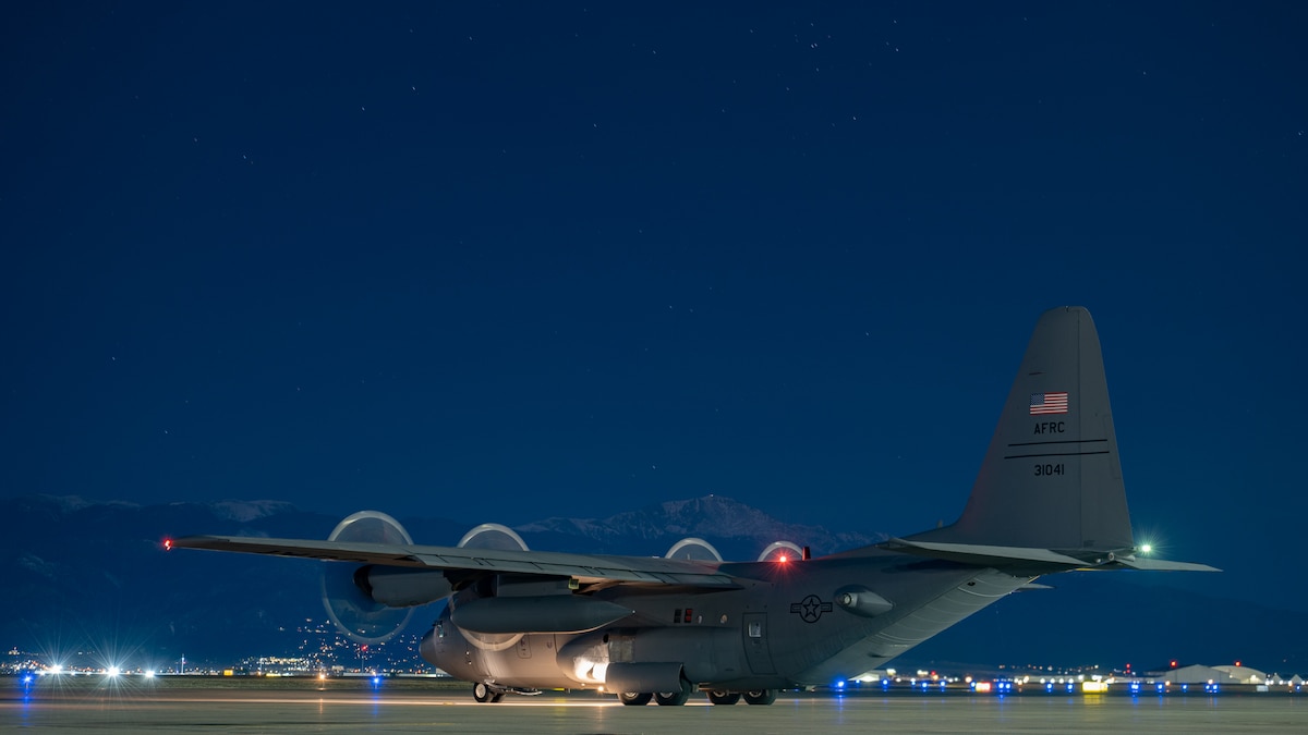 A military cargo aircraft on the parking ramp at night with a mountaintop in the background.