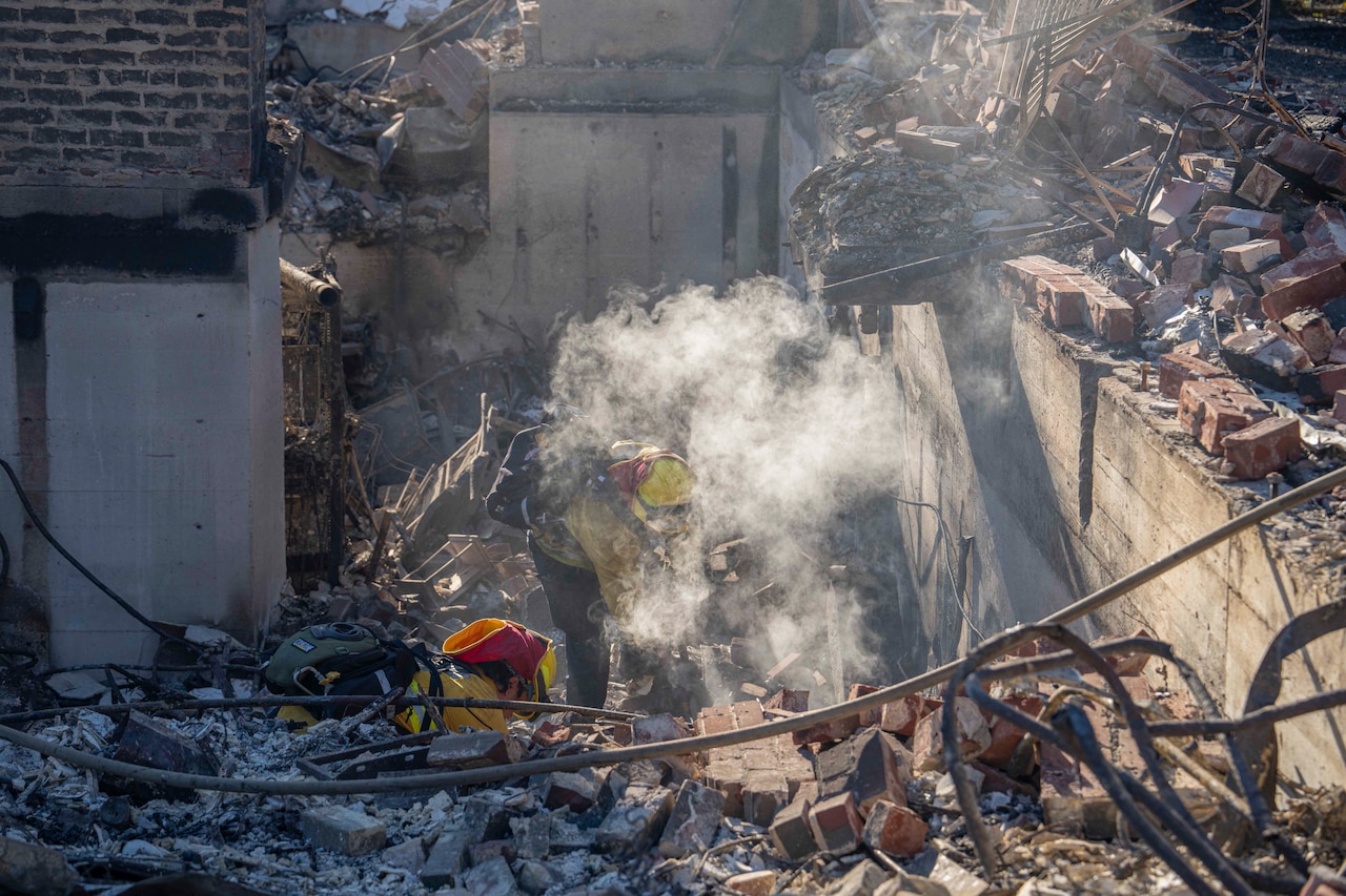 Two firefighters work in the ruins of a building after a wildfire during the day.