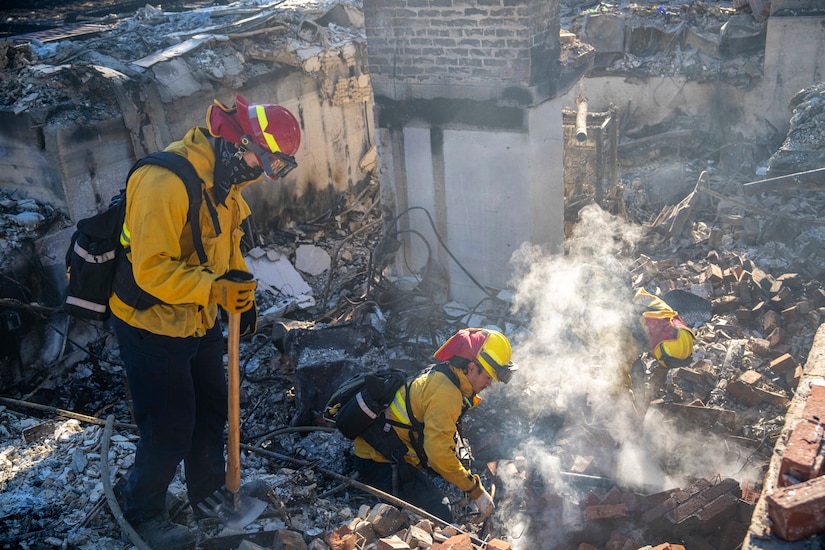 Two firefighters in yellow gear sift through the rubble of a burned out building while a third observes from nearby.