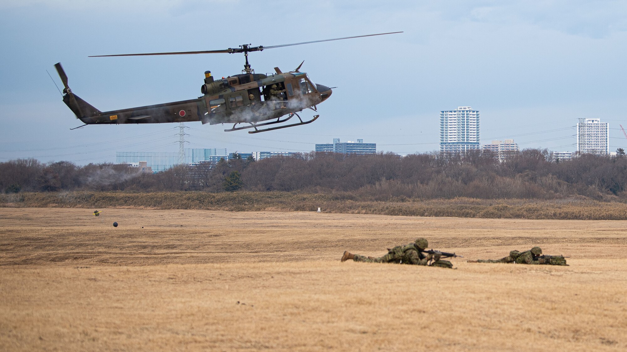A Huey helicopter fires blank rounds while flying over two soldiers