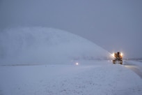 Snow flies in the air as an airman operates a snow blower on a flight line illuminated by yellowish spotlights.