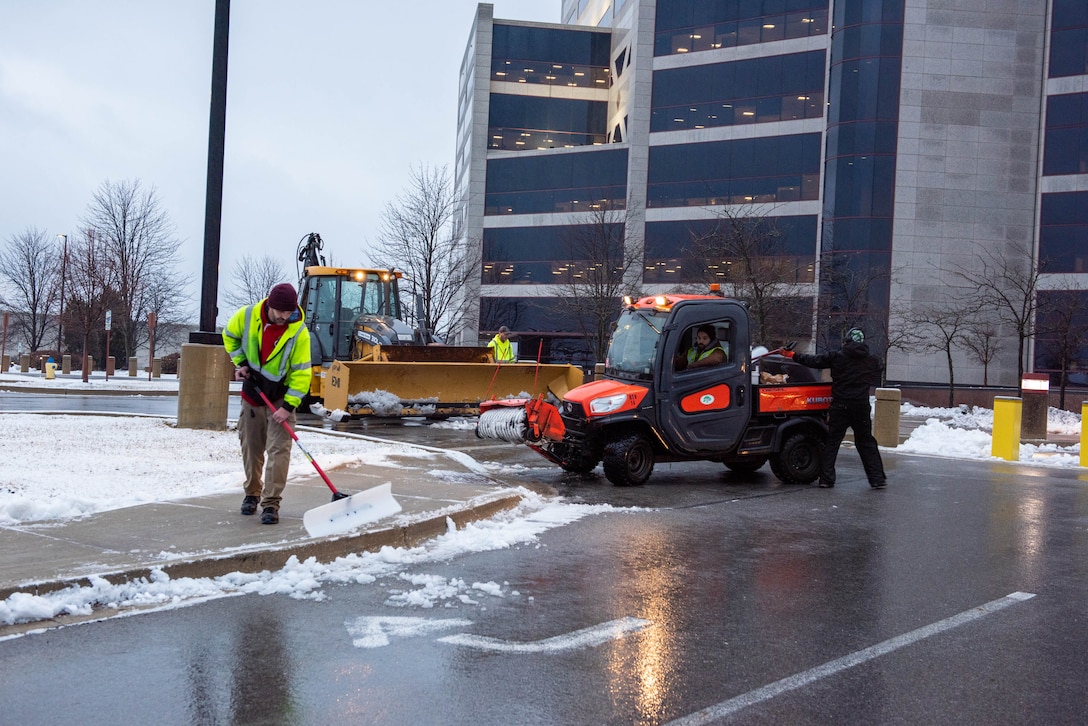 A man in a safety vest shovels snow off sidewalks as a man with a plow cleans roads in front of a building.
