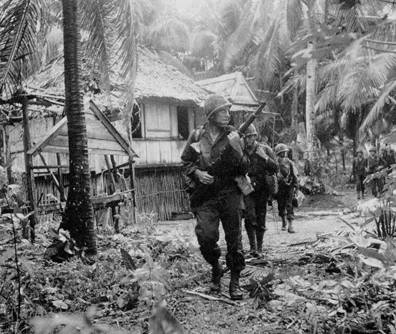 Three men in combat uniforms walk through a jungle area near a thatched building.
