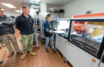 Rear Adm. Mark Sucato, commander, Navy Region Northwest and Chad Davis, business activity director, Morale Welfare and Recreation, Naval Base Kitsap-Bremerton, watch as Joyce Embree, production lead, MWR, demonstrates how the Picnic Pizza Station, a new automated pizza machine, makes pizza, Dec. 9, 2024, inside the Building 435 cafeteria at Puget Sound Naval Shipyard & Intermediate Maintenance Facility in Bremerton, Washington. (U.S. Navy photo by Wendy Hallmark)