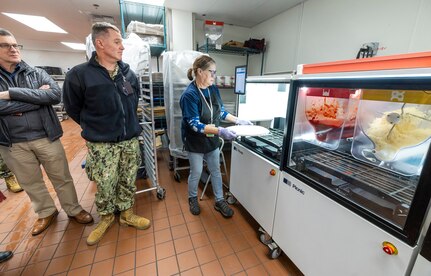 Rear Adm. Mark Sucato, commander, Navy Region Northwest and Chad Davis, business activity director, Morale Welfare and Recreation, Naval Base Kitsap-Bremerton, watch as Joyce Embree, production lead, MWR, demonstrates how the Picnic Pizza Station, a new automated pizza machine, makes pizza, Dec. 9, 2024, inside the Building 435 cafeteria at Puget Sound Naval Shipyard & Intermediate Maintenance Facility in Bremerton, Washington. (U.S. Navy photo by Wendy Hallmark)