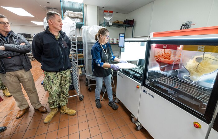 Rear Adm. Mark Sucato, commander, Navy Region Northwest and Chad Davis, business activity director, Morale Welfare and Recreation, Naval Base Kitsap-Bremerton, watch as Joyce Embree, production lead, MWR, demonstrates how the Picnic Pizza Station, a new automated pizza machine, makes pizza, Dec. 9, 2024, inside the Building 435 cafeteria at Puget Sound Naval Shipyard & Intermediate Maintenance Facility in Bremerton, Washington. (U.S. Navy photo by Wendy Hallmark)