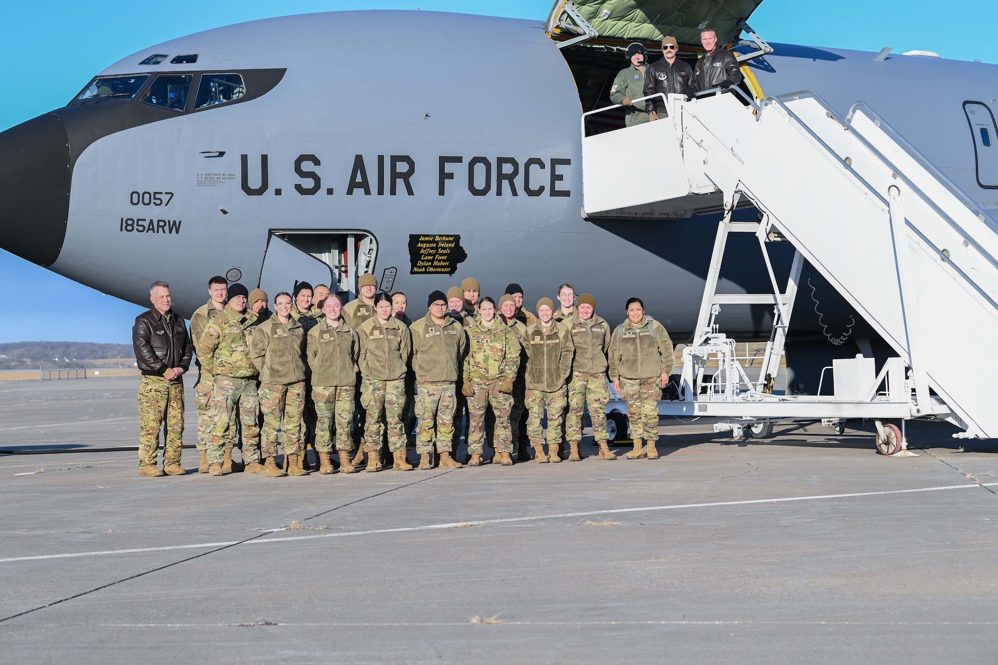 A group of U.S. soldiers and Airmen wearing camouflage uniforms stand as a group with a large grey Air Force aircraft in background