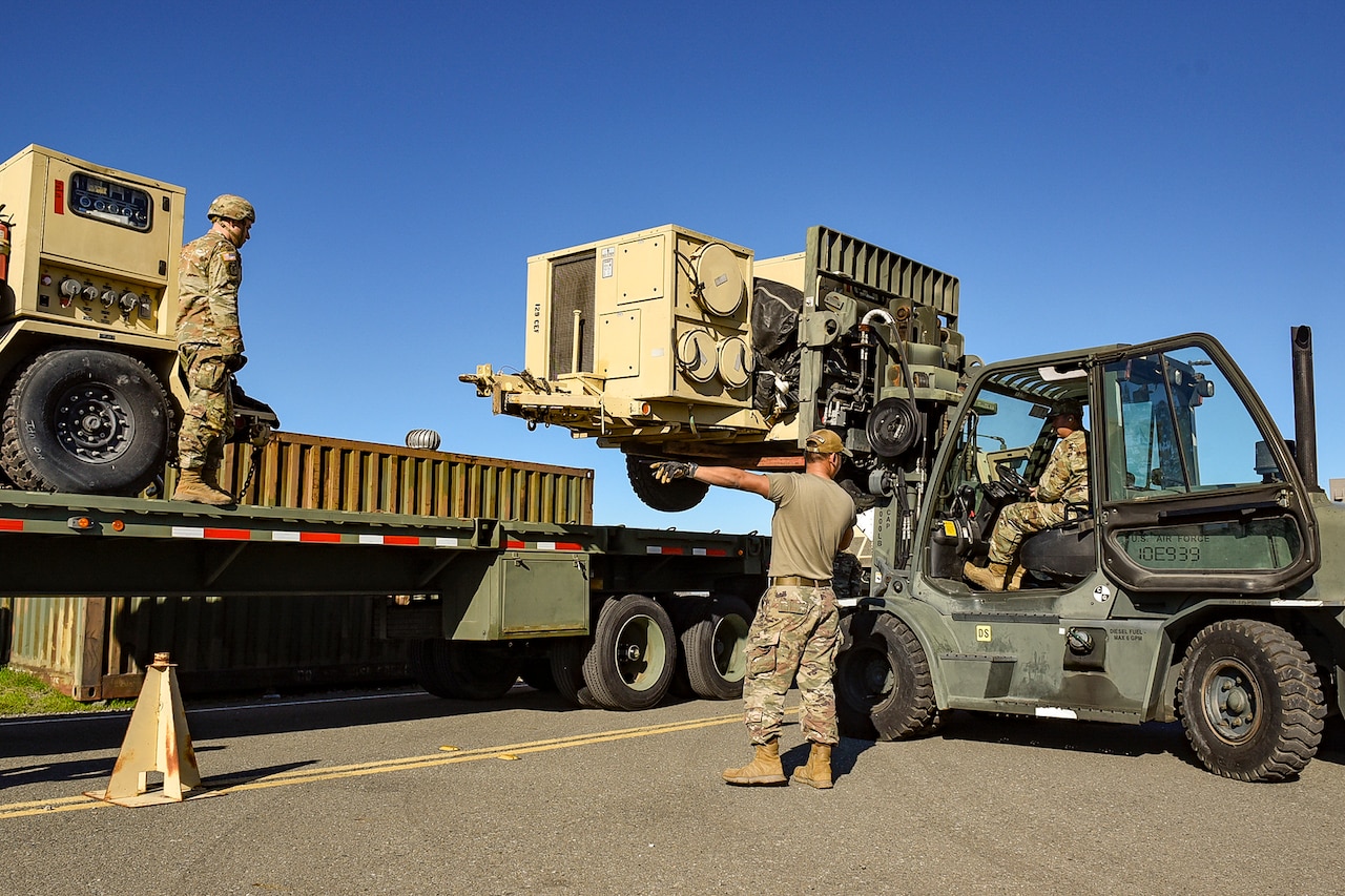 A group of guardsmen load a pallet onto the back of a truck during a clear day.