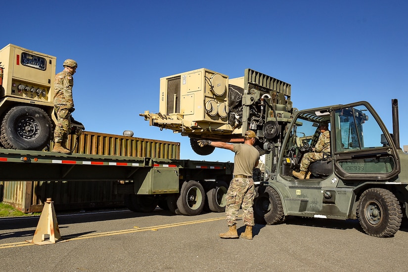 A group of guardsmen load a pallet onto the back of a truck during a clear day.