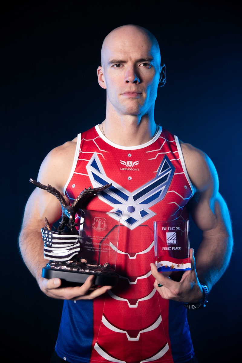 Tim Gribble poses for a portrait wearing a red and blue tank top with the U.S. Air Force logo while holding two trophies.
