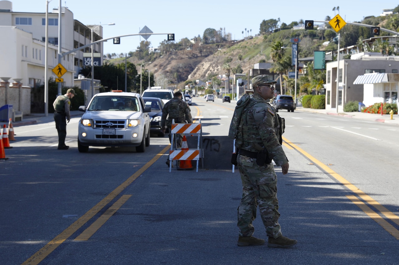 A soldier monitors a road during a sunny day with other soldiers walking around and talking to people in cars.