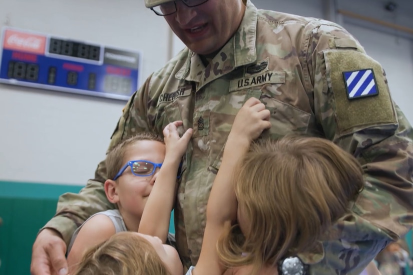 Three kids hug a soldier in uniform in a gym-type room.