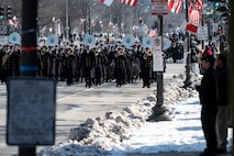 Service members march with pedestrians lining the sidewalks to watch