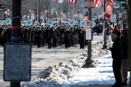 Service members march with pedestrians lining the sidewalks to watch