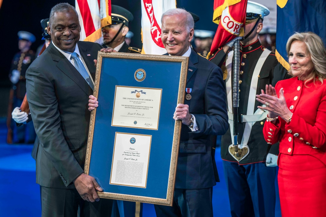 Two men hold a large frame that contains two certificates. Behind them are soldiers.