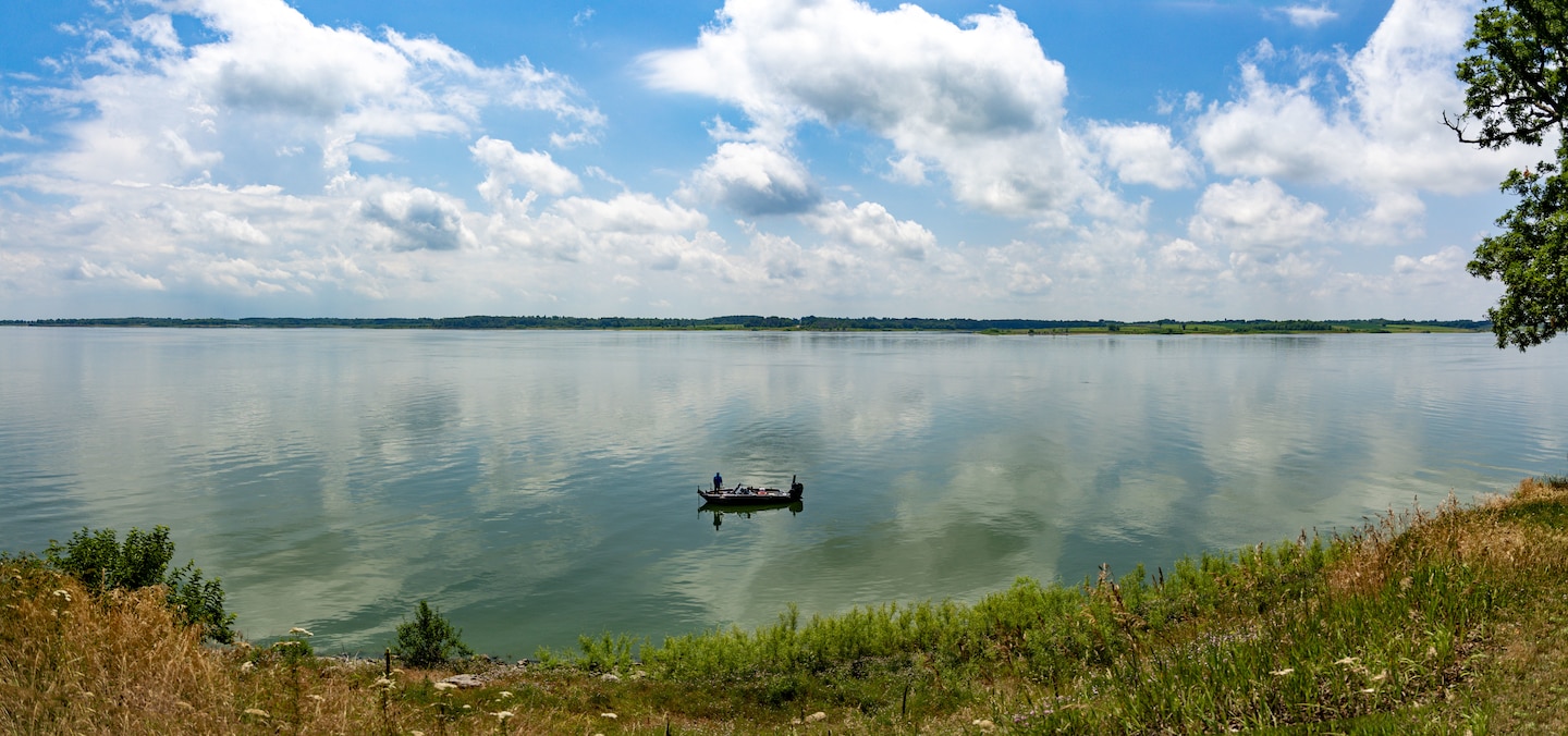 A body of water with a cloudy sky in the background and brown vegetation in the foreground.