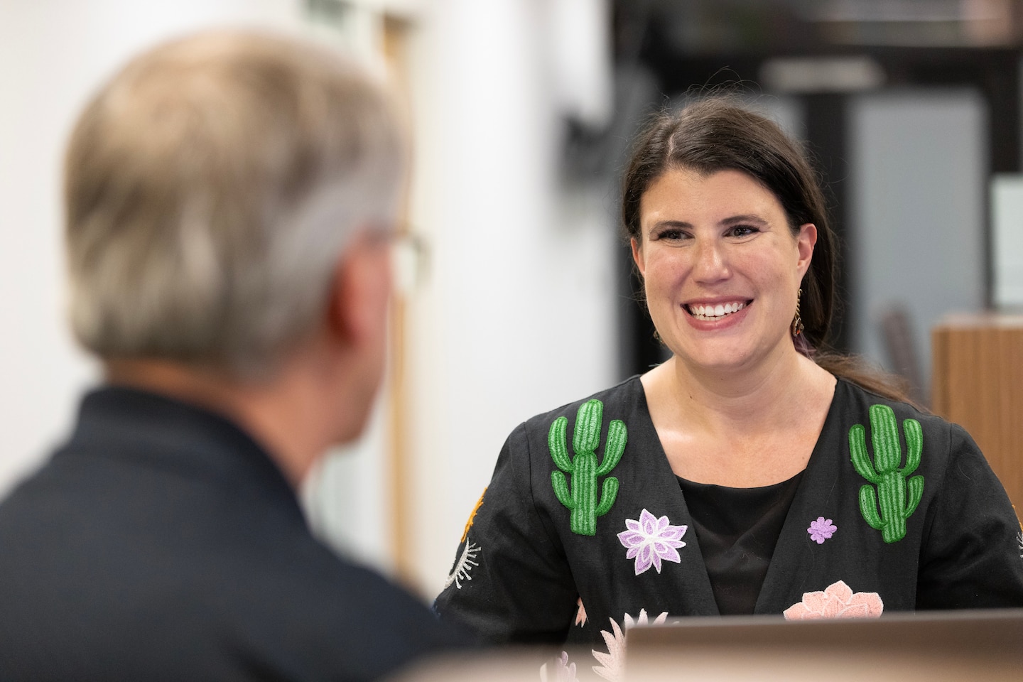 A man in a black shirt has his back turned and a woman in a black shirt with green cacti on the shoulders looks at him and smiles.
