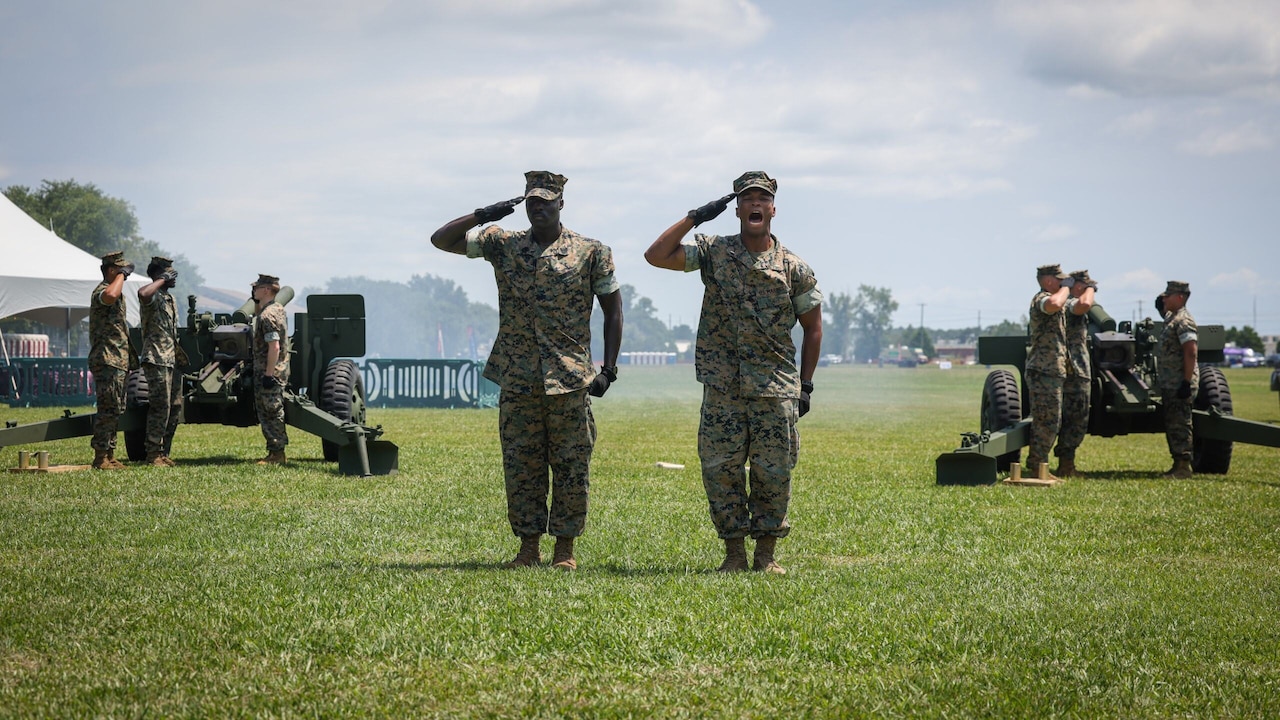 Two men salute in a field as two crews shoot ceremonial howitzer shots into the distance.