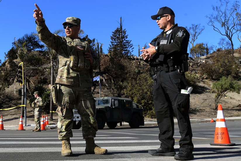 A solider standing in a street points while talking to a police officer.