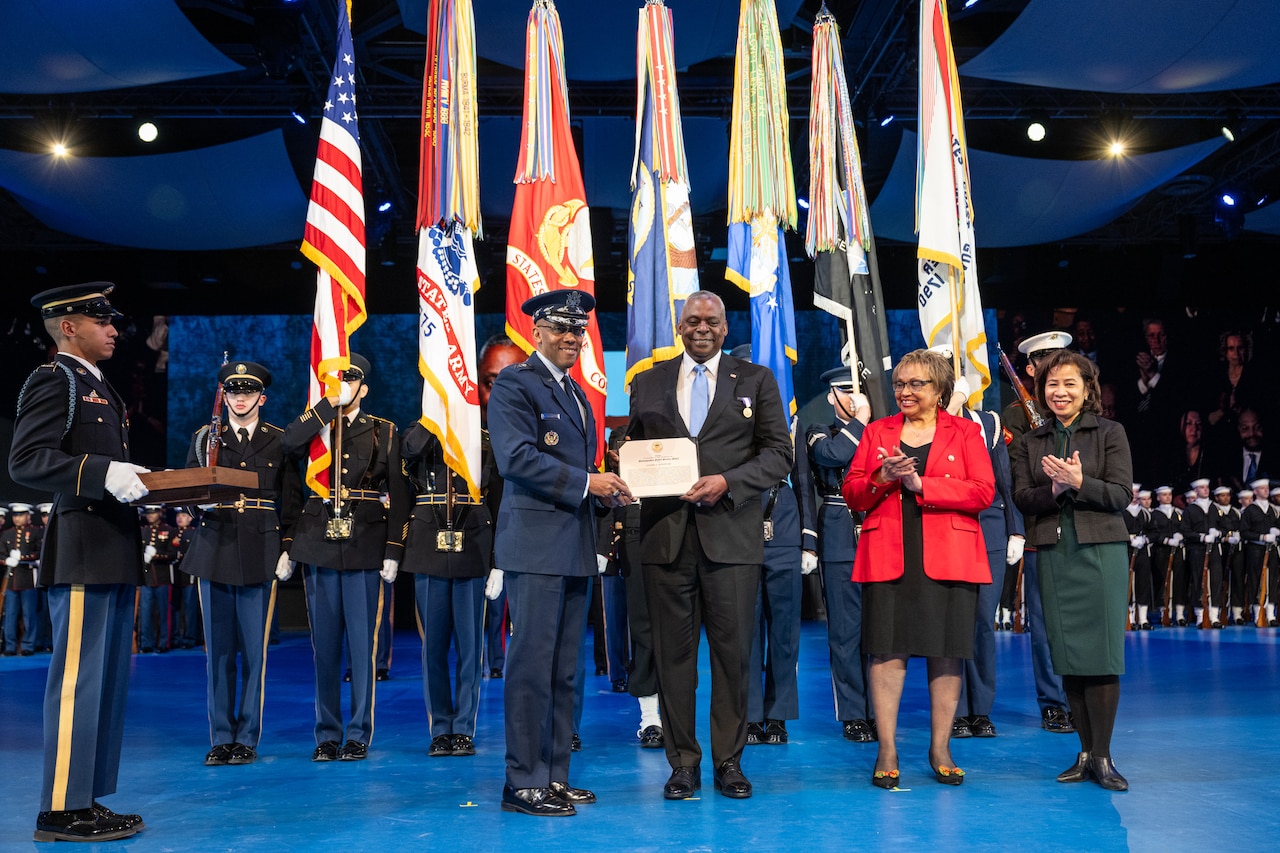 Two men smile while holding a plaque in front of troops holding flags during a ceremony.