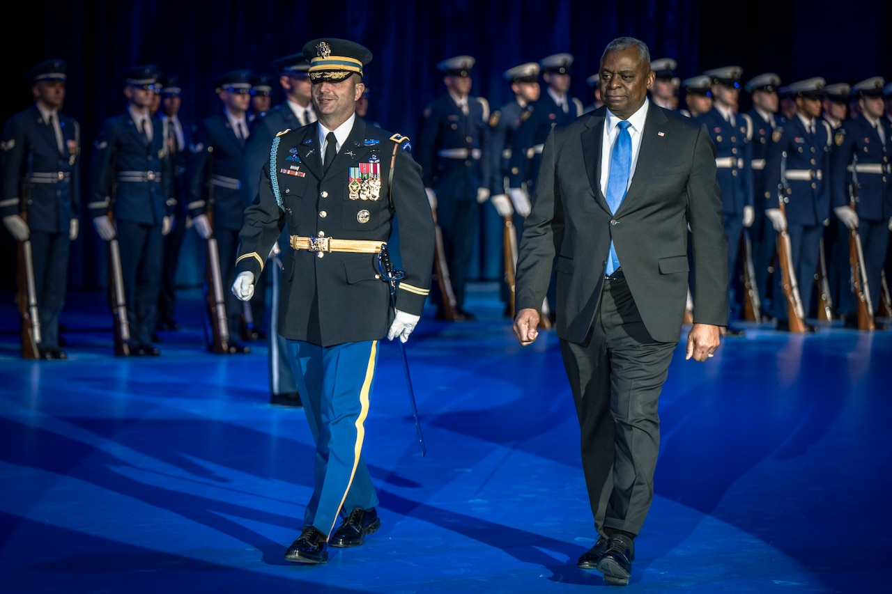 A man in a suit walks along aside a man in uniform on stage. Men in uniform holding rifles stand in the background.
