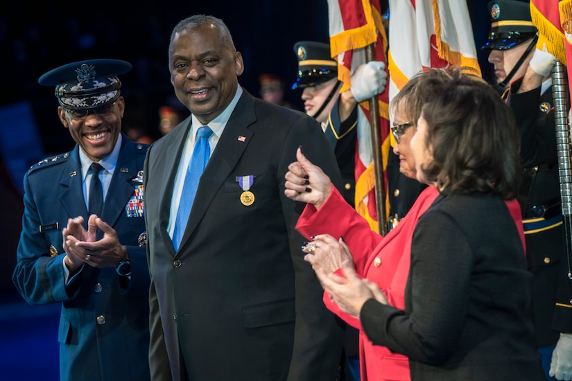 A man in a suit with a medal smiles as on lookers clap for him and give him thumbs up.