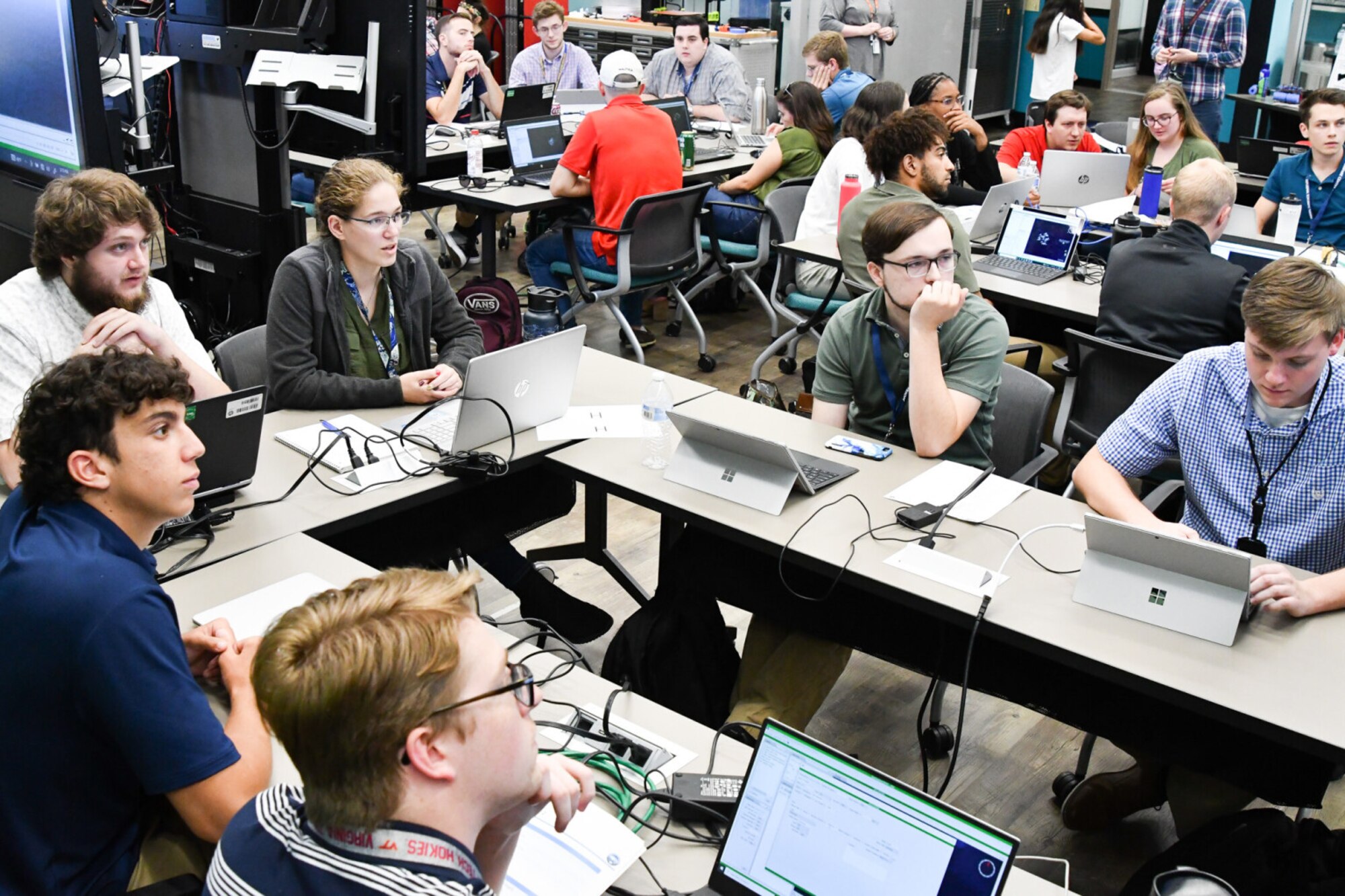 A group of students sit at a table and watch a monitor.