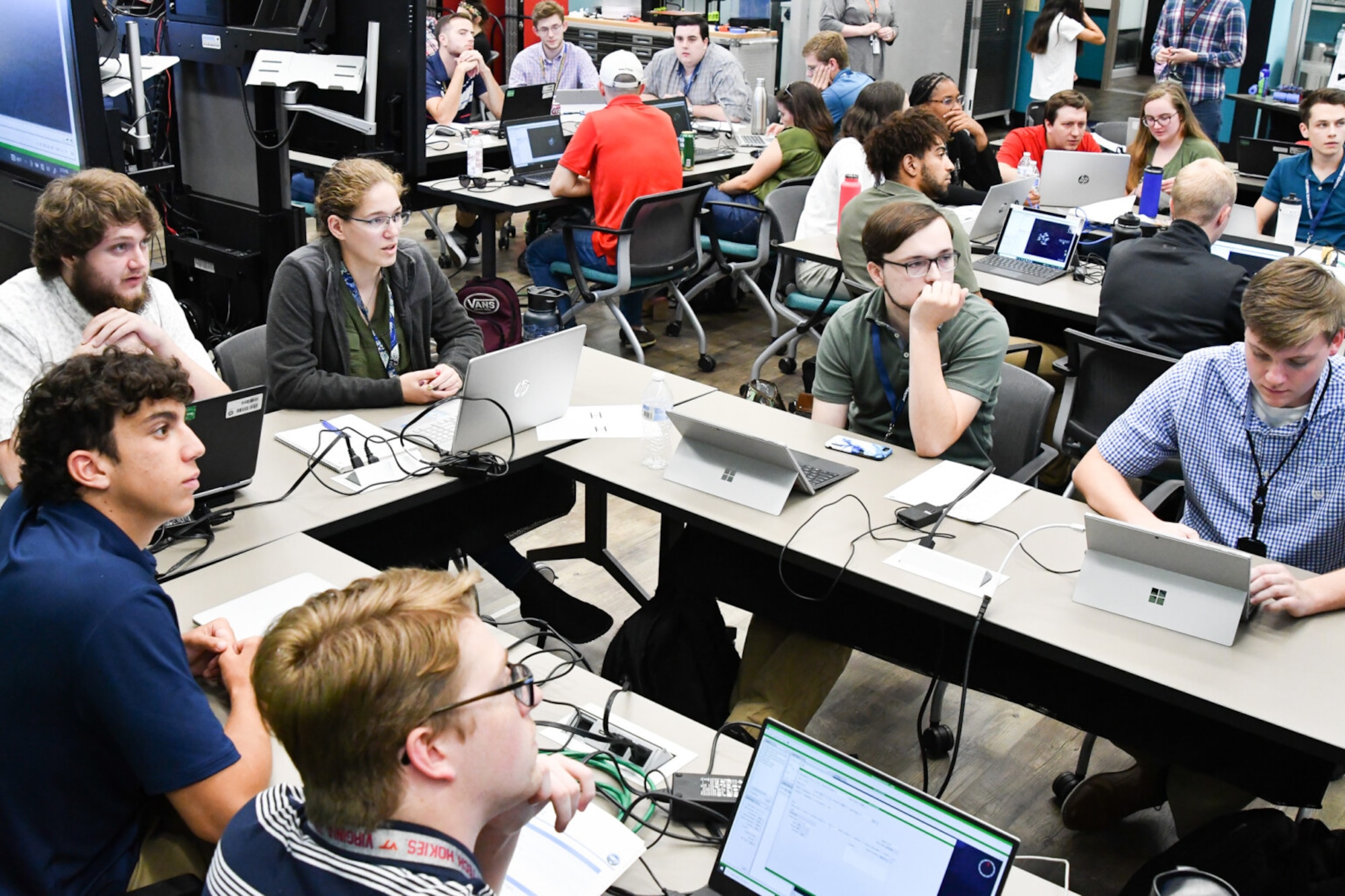 A group of students sit at a table and watch a monitor.