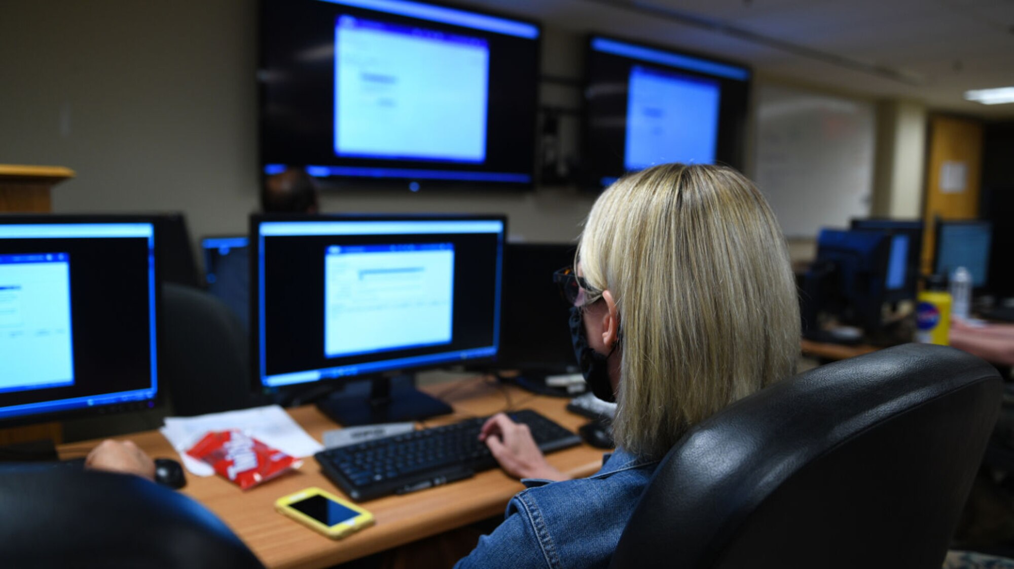 A woman, facing away, types at a keyboard and watches two monitors.