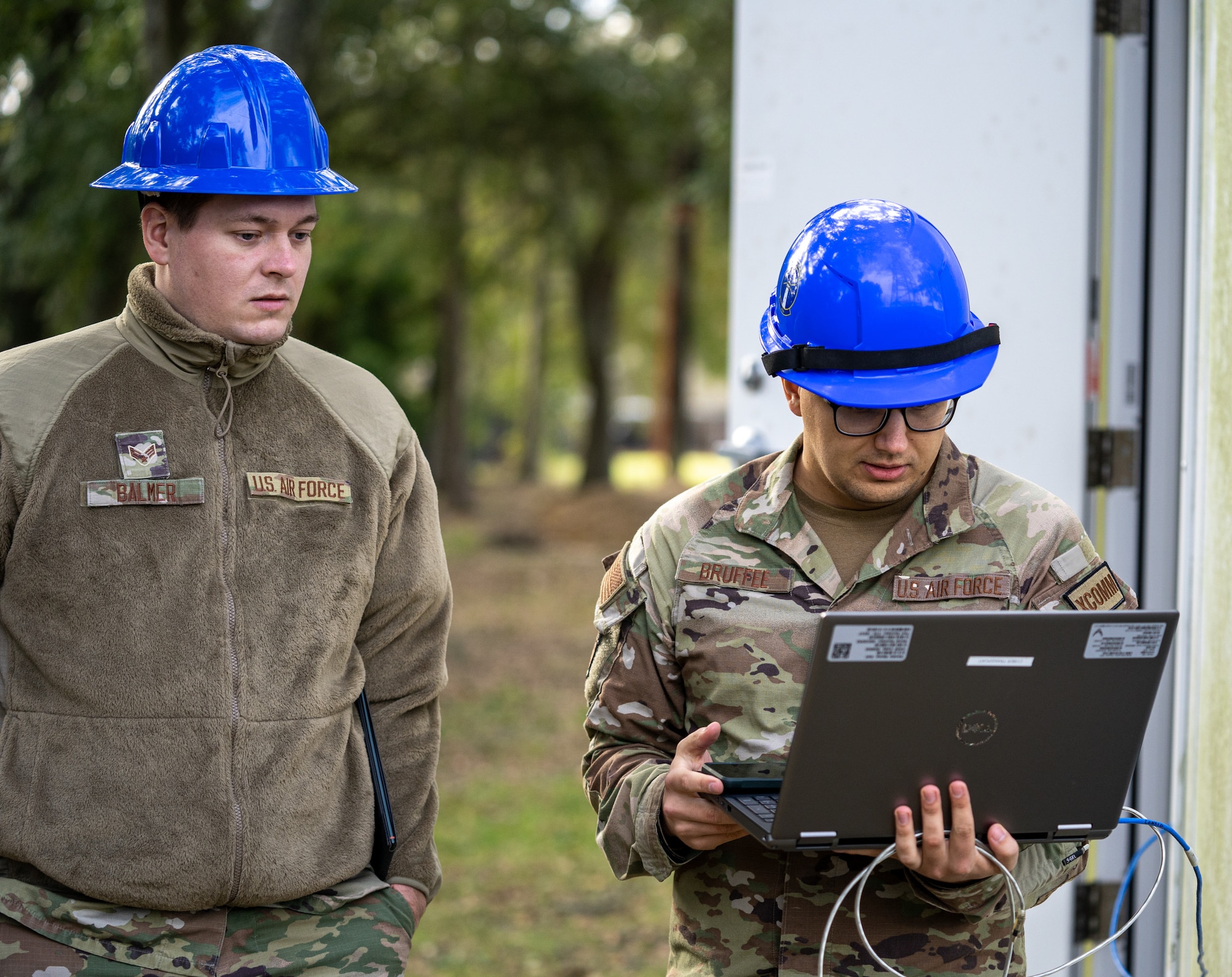 One service man in an operational camouflage pattern uniform stands behind another who is holding a laptop.