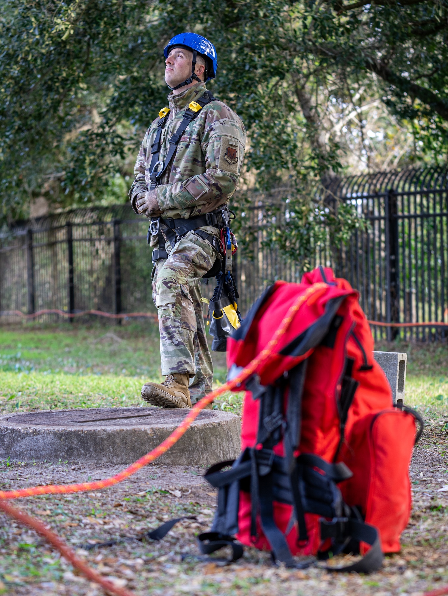 A serviceman in an operational camouflage pattern uniform wearing a blue hardhat and tower climbing gear looks up from the ground while a red backpack sits beside him out of focus.