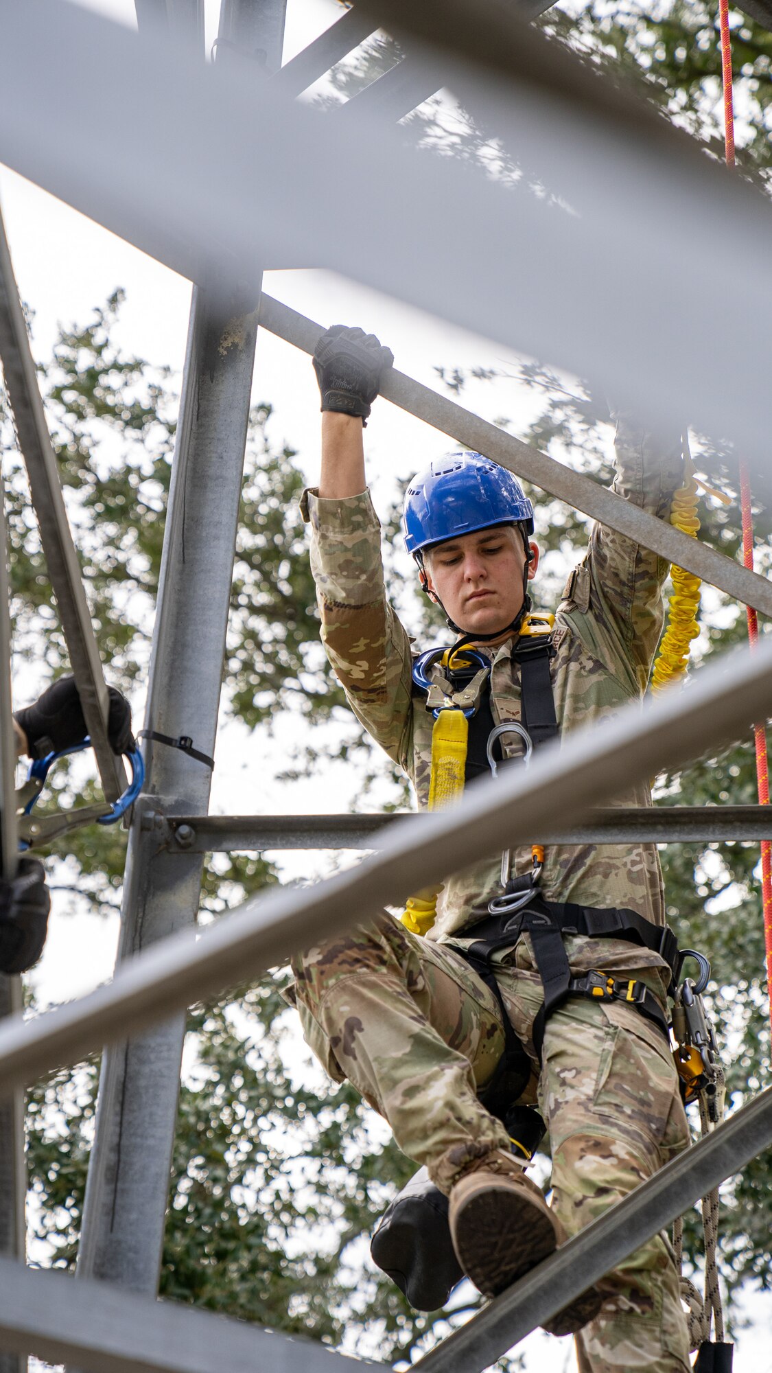 A serviceman in an operational camouflage pattern uniform wearing a blue hardhat and tower climbing harness scales a tower.