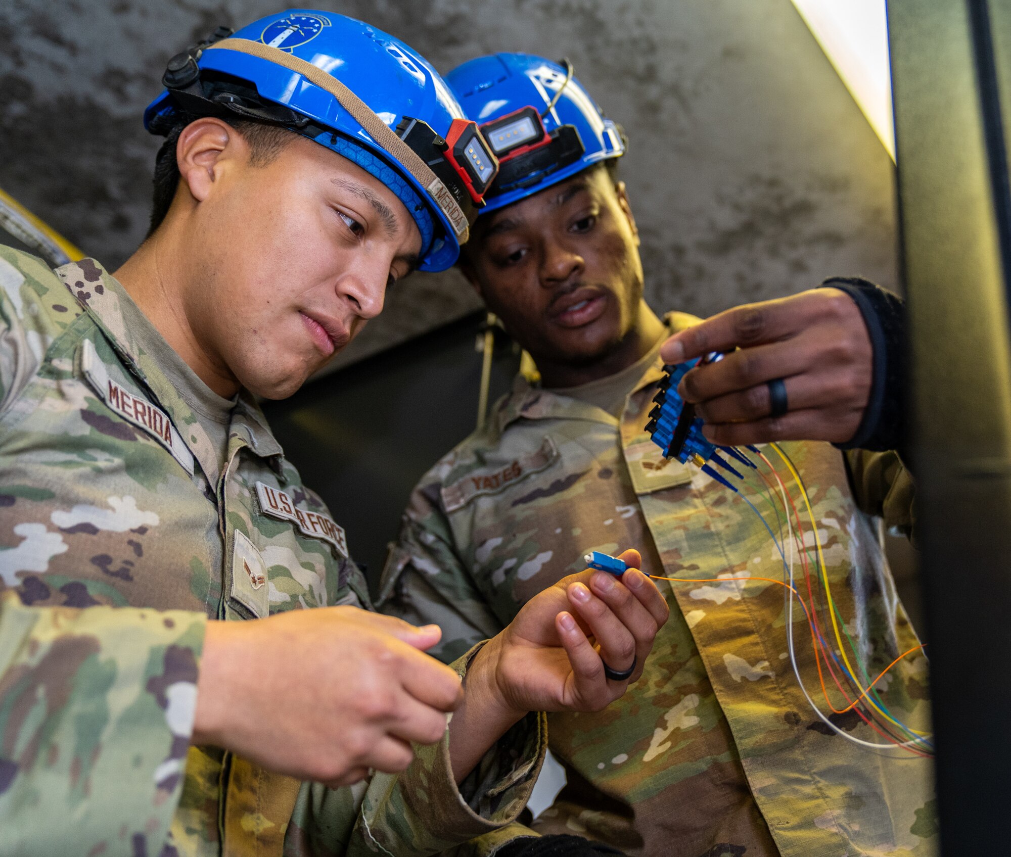 Two servicemen in operational camouflage pattern uniforms wearing blue hardhats inspect wires.