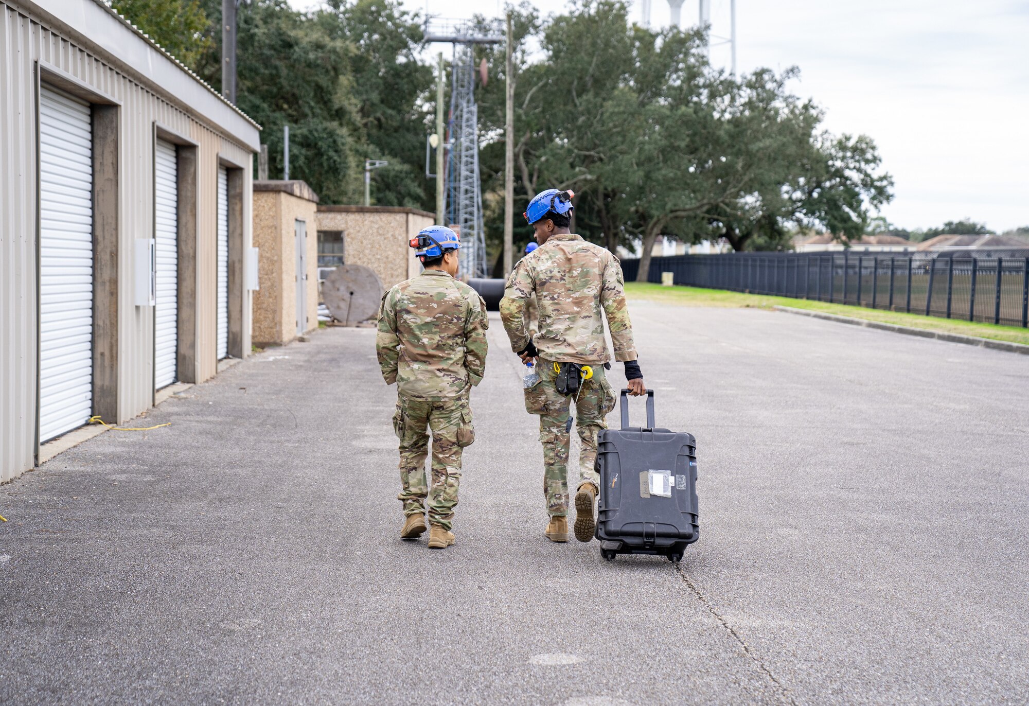 Two servicemen in operational camouflage pattern uniforms talk to each other while rolling a black box with wheels behind them.